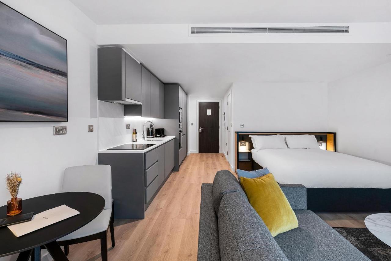 Citysuites 2 Aparthotel Manchester Exteriér fotografie The photo shows a modern and stylish studio apartment. It features a kitchenette with dark cabinetry and a small dining table with two chairs. There's a cozy sofa with colorful cushions, positioned next to the table. In the background, a large bed wi