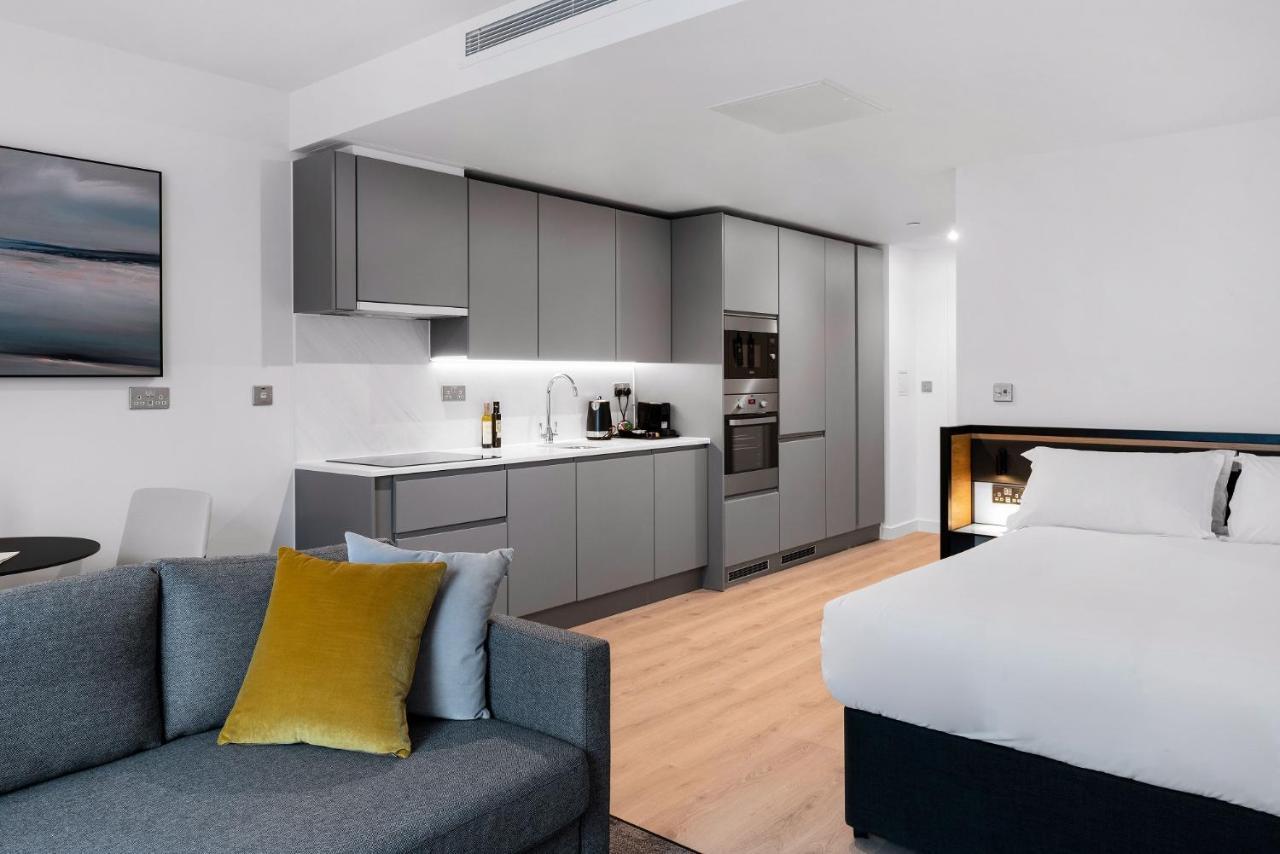 Citysuites 2 Aparthotel Manchester Exteriér fotografie The photo shows a modern studio apartment featuring an open-plan layout. On one side, there is a kitchenette with sleek, gray cabinets and appliances, including a coffee maker and oven. Adjacent to the kitchenette is a small dining area with a table 