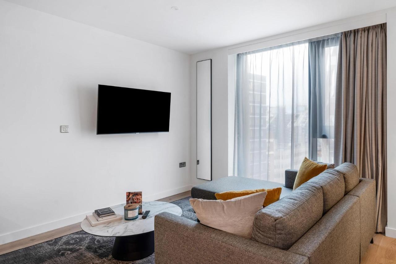 Citysuites 2 Aparthotel Manchester Exteriér fotografie The image shows a modern living room with a minimalist design. There is a large flat-screen TV mounted on the white wall. A comfortable gray sofa with textured cushions occupies the center of the room, complemented by a round coffee table in front of