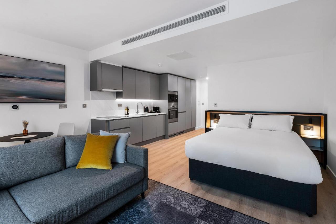 Citysuites 2 Aparthotel Manchester Exteriér fotografie The photo shows a modern studio apartment. On one side, there is a comfortable gray sofa adorned with a yellow cushion. Nearby, a round table is placed, likely intended for dining or casual sitting. 

The kitchen area features sleek gray cabinetry wi