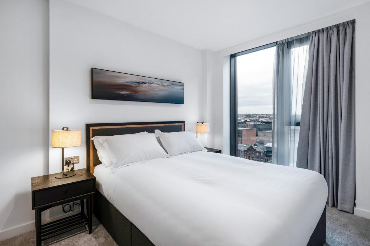 Citysuites 2 Aparthotel Manchester Exteriér fotografie The image shows a modern bedroom featuring a large bed with a white duvet and two pillows. On either side of the bed are small nightstands with lamps. A large window occupies one wall, draped with light gray curtains, allowing natural light to enter 