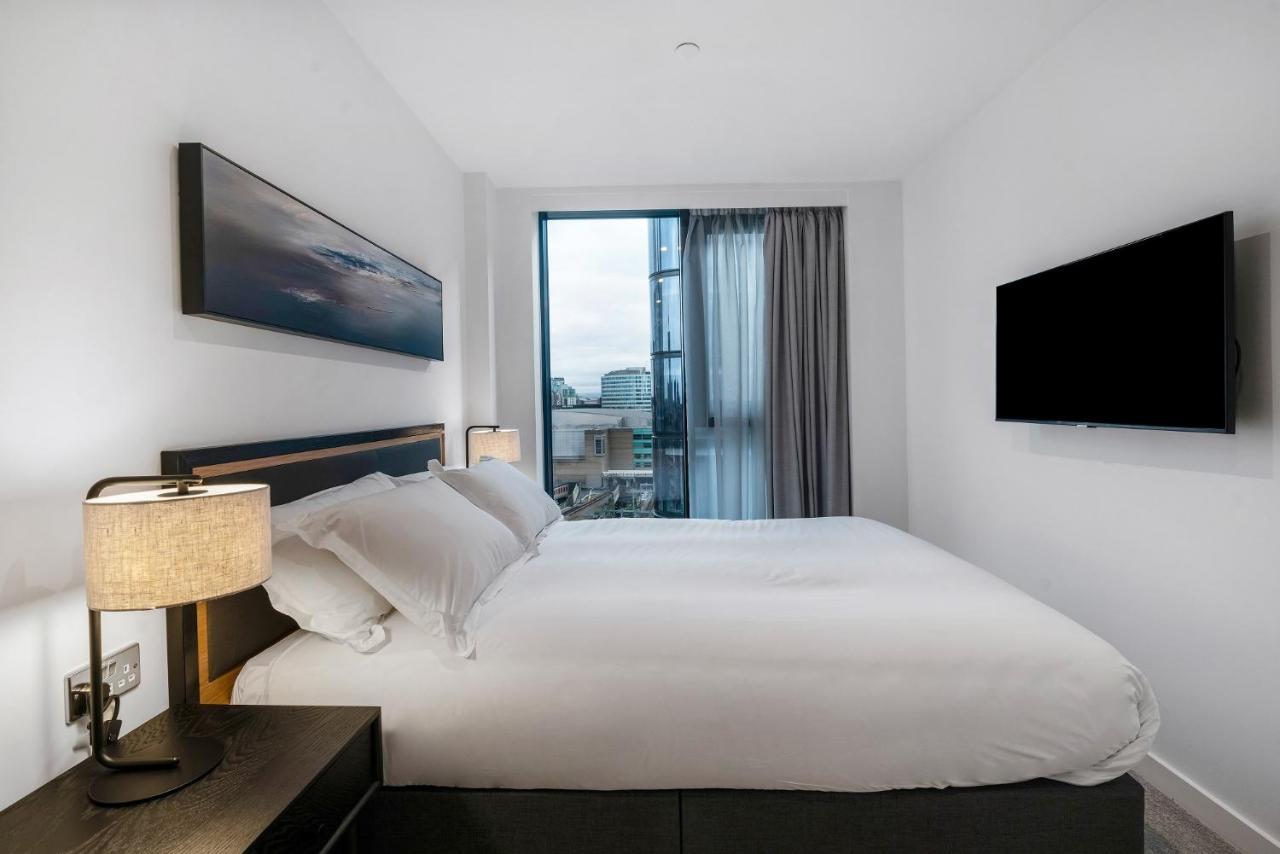 Citysuites 2 Aparthotel Manchester Exteriér fotografie The photo shows a modern bedroom with a minimalist aesthetic. It features a large bed with white bedding and two pillows. On either side of the bed, there are bedside tables with lamps. A large window lets in natural light, covered by sheer curtains.