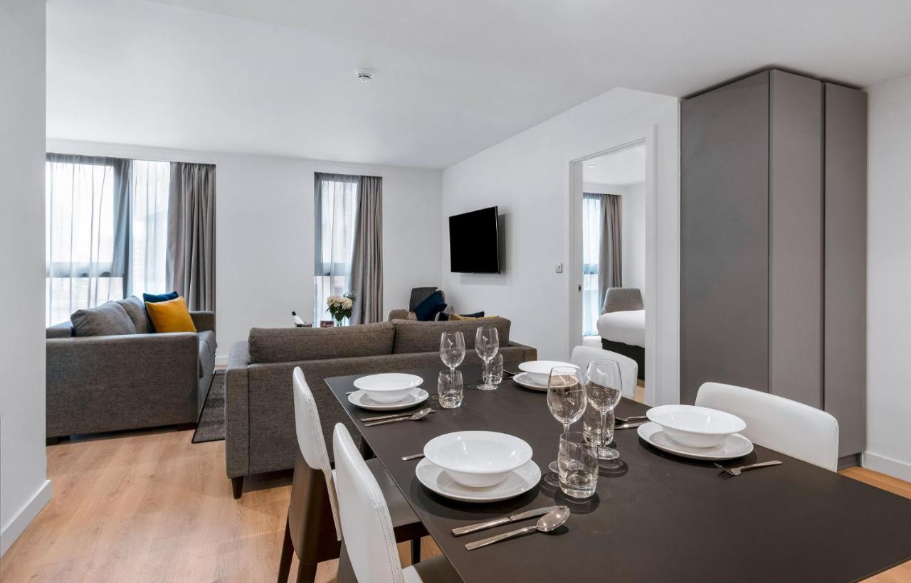 Citysuites 2 Aparthotel Manchester Exteriér fotografie The photo shows a modern living space with an open layout. In the foreground, there is a dining table set for a meal, featuring multiple plates, glasses, and cutlery arranged neatly. In the background, there is a comfortable-looking gray sofa with a 