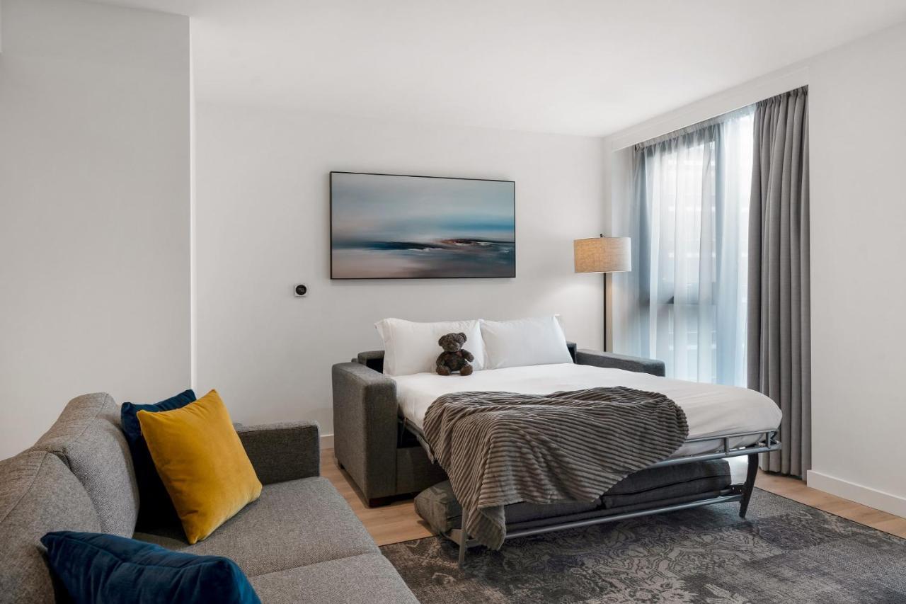 Citysuites 2 Aparthotel Manchester Exteriér fotografie The photo shows a modern and minimalistic guest room. There is a sofa on the left side, adorned with a colorful cushion. In the middle is a bed that appears to be a pull-out style, featuring white bedding and a gray throw blanket. A stuffed toy is pl