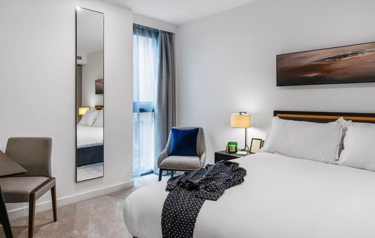 Citysuites 2 Aparthotel Manchester Exteriér fotografie The photo shows a modern hotel room. It features a neatly made bed with white linens and a dark-colored throw draped over it. There is a small bedside table with a lamp next to the bed. A comfortable chair in a light color is positioned near the wind