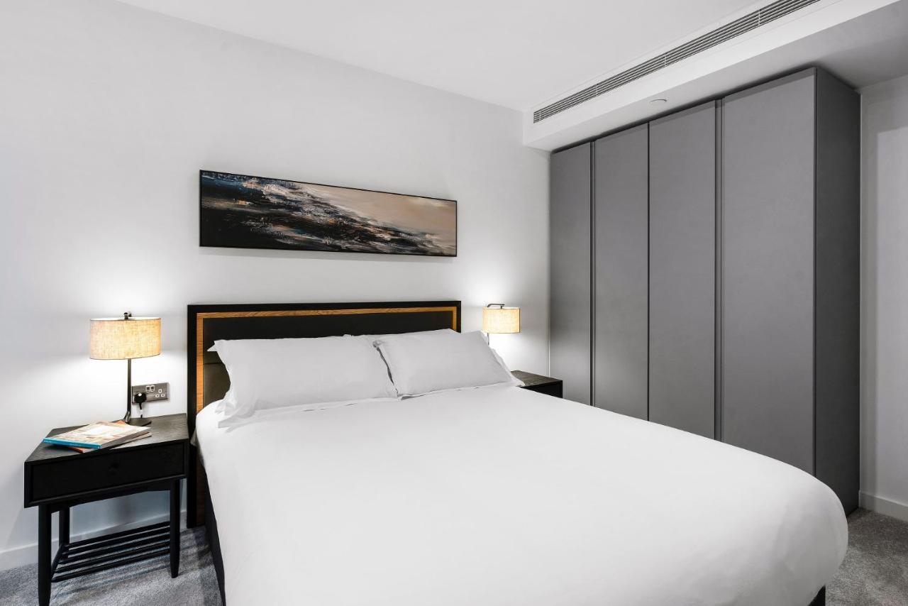 Citysuites 2 Aparthotel Manchester Exteriér fotografie The photo shows a modern bedroom designed with a minimalist aesthetic. The focal point is a large bed with white bedding, positioned against a wall. On either side of the bed, there are black nightstands topped with lamps, providing a warm glow. Abov