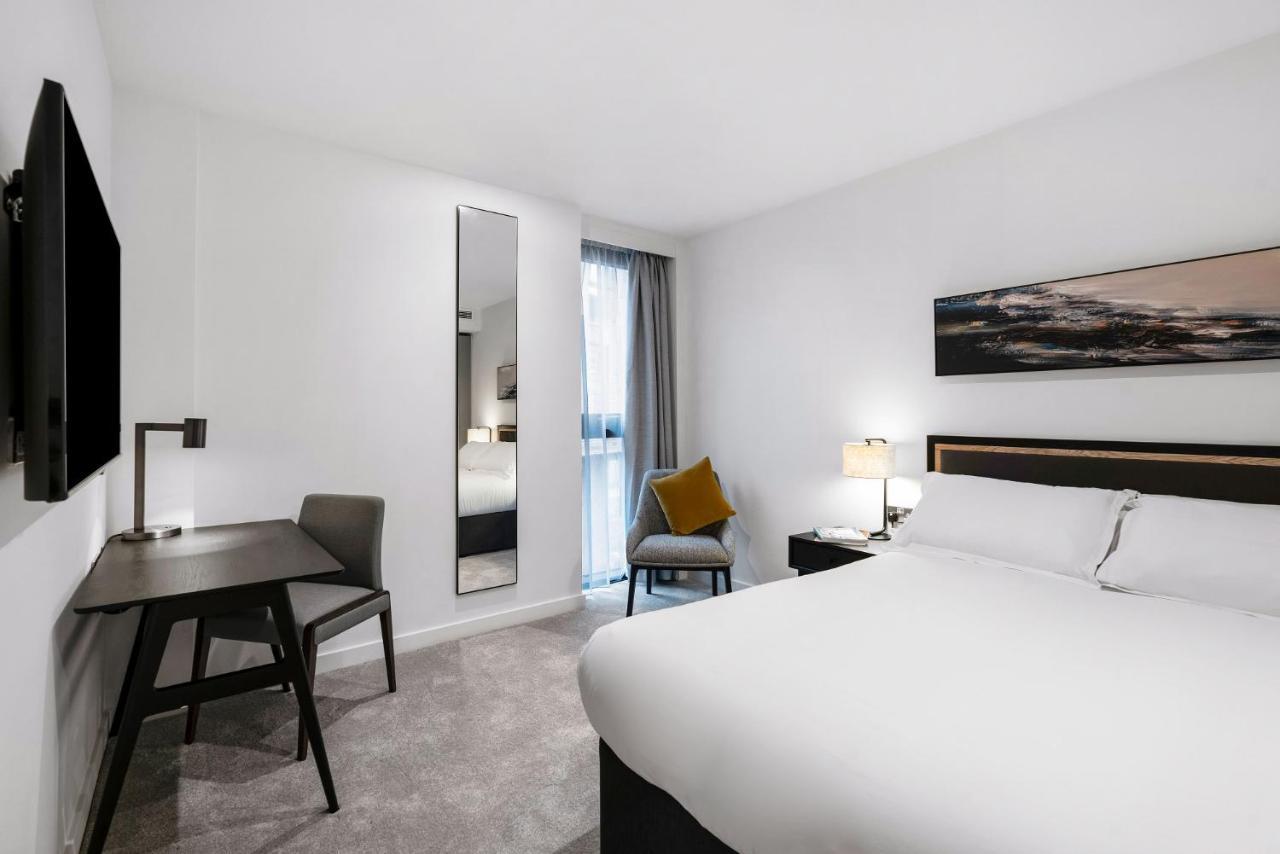 Citysuites 2 Aparthotel Manchester Exteriér fotografie The photo shows a modern hotel room with a clean and minimalist design. It features a large bed with white linens, a small desk with a chair, and a mirror mounted on the wall. There is a chair in one corner with a decorative pillow, and the room has 