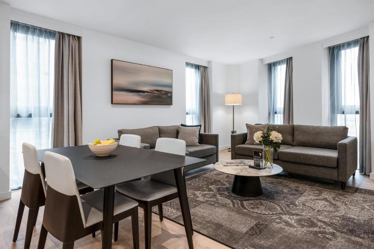 Citysuites 2 Aparthotel Manchester Exteriér fotografie The photo depicts a modern and stylish interior of a living space. On the left, there is a dining table with four chairs, featuring a bowl of fruits on the table. In the background, there's a cozy seating area with a gray sofa and a smaller round cof