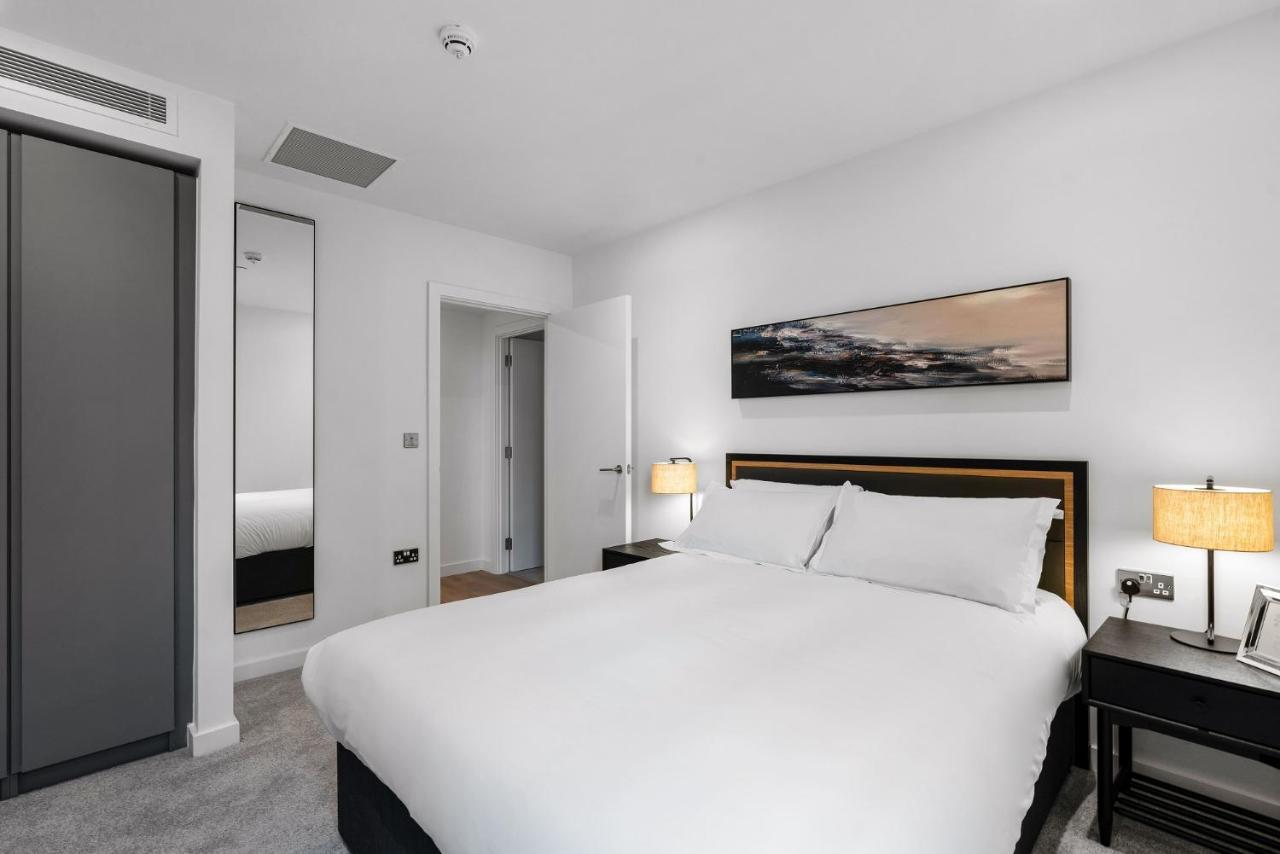 Citysuites 2 Aparthotel Manchester Exteriér fotografie The photo shows a modern bedroom featuring a large double bed with white bedding. There are two bedside tables with lamps on either side of the bed. The walls are painted white, and there's a piece of artwork above the bed that depicts a landscape. A