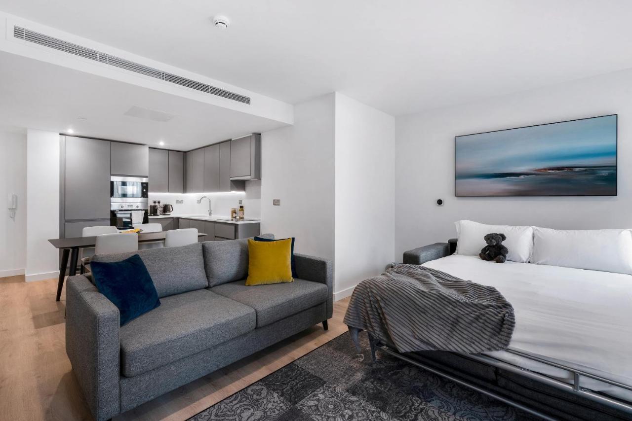 Citysuites 2 Aparthotel Manchester Exteriér fotografie The photo shows a modern, open-concept living space that includes a kitchen and a sleeping area. 

- **Living Area**: On the left, there's a gray sofa with a blue and yellow throw pillow. A blanket is draped over one side, and there's a small dark ru
