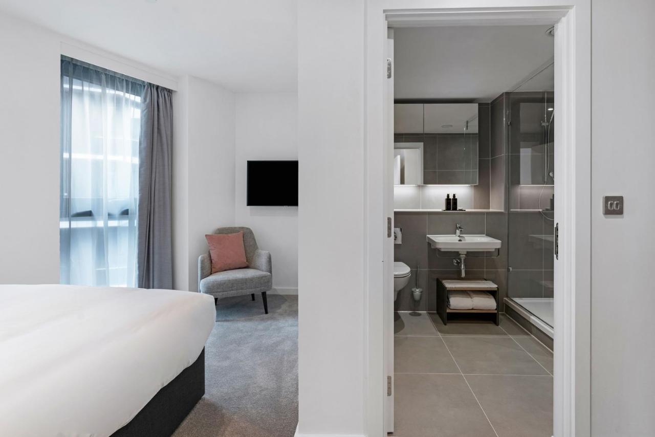 Citysuites 2 Aparthotel Manchester Exteriér fotografie The photo shows a modern hotel room and bathroom setup. On the left, there is a bed with white bedding and a small sitting area that includes a gray chair and a pillow in a subtle pink shade. A flat-screen TV is mounted on the wall, providing enterta