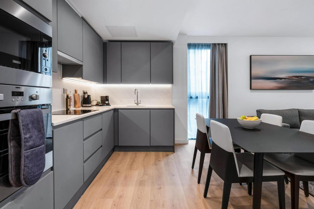 Citysuites 2 Aparthotel Manchester Exteriér fotografie The photo shows a modern kitchen and dining area. The kitchen features sleek gray cabinetry, a white countertop, and built-in appliances such as an oven. There is a sink and some kitchen utensils displayed. The adjacent dining area contains a black t