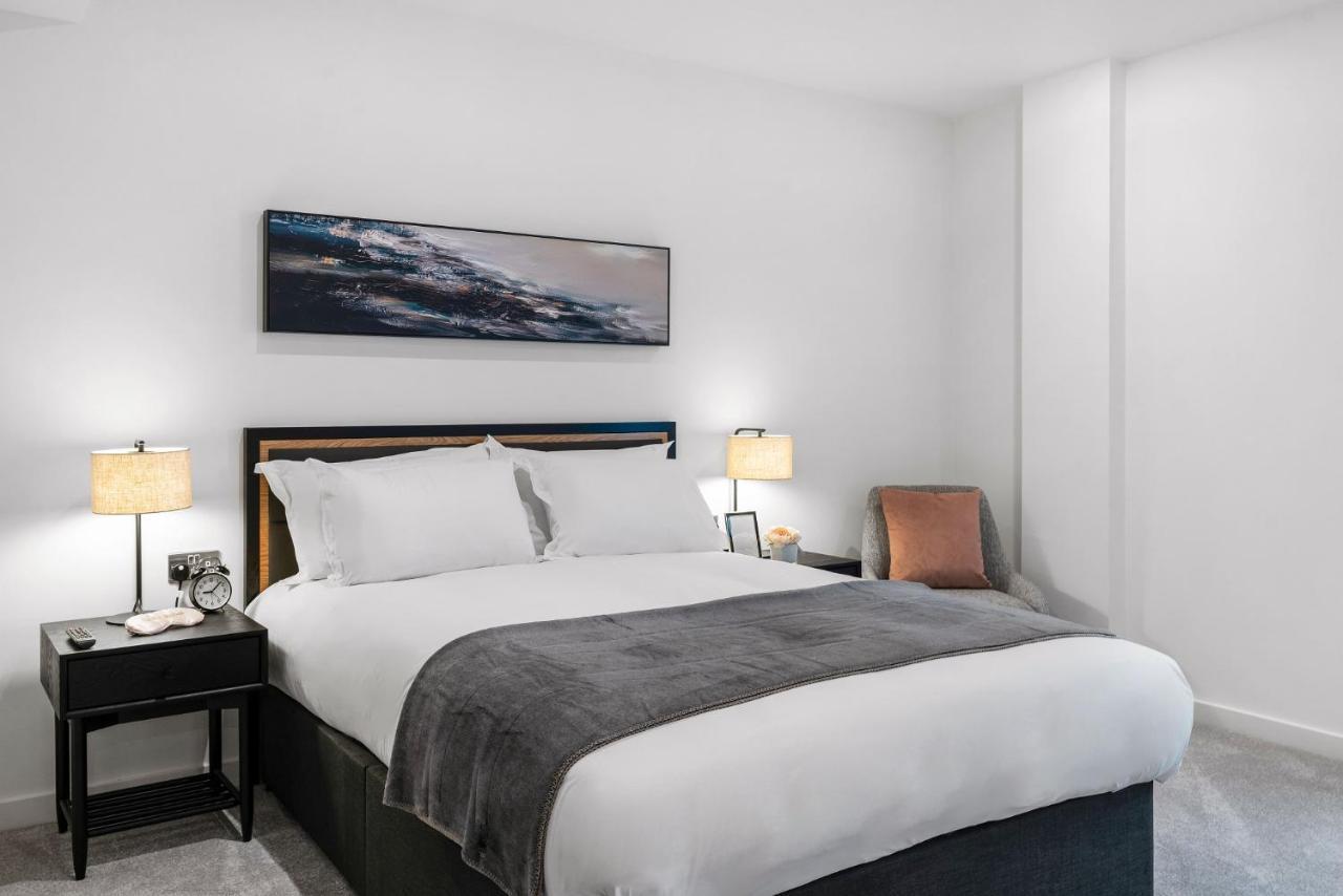 Citysuites 2 Aparthotel Manchester Exteriér fotografie The photo shows a modern bedroom featuring a large bed with crisp white linens and a grey throw blanket. On either side of the bed, there are bedside tables with lamps. A piece of abstract art hangs above the headboard, and there is a chair in a corn