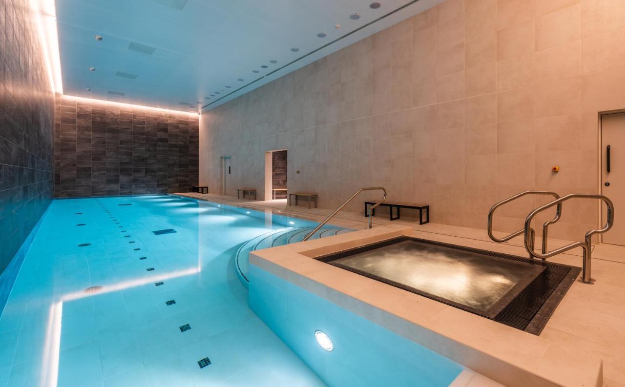 Citysuites 2 Aparthotel Manchester Exteriér fotografie The photo shows an indoor swimming pool area. The pool is a bright blue color and features smooth, tiled surfaces. On one side, there is a hot tub area with steps leading into it. The walls are made of light-colored stone, and there is ambient lighti