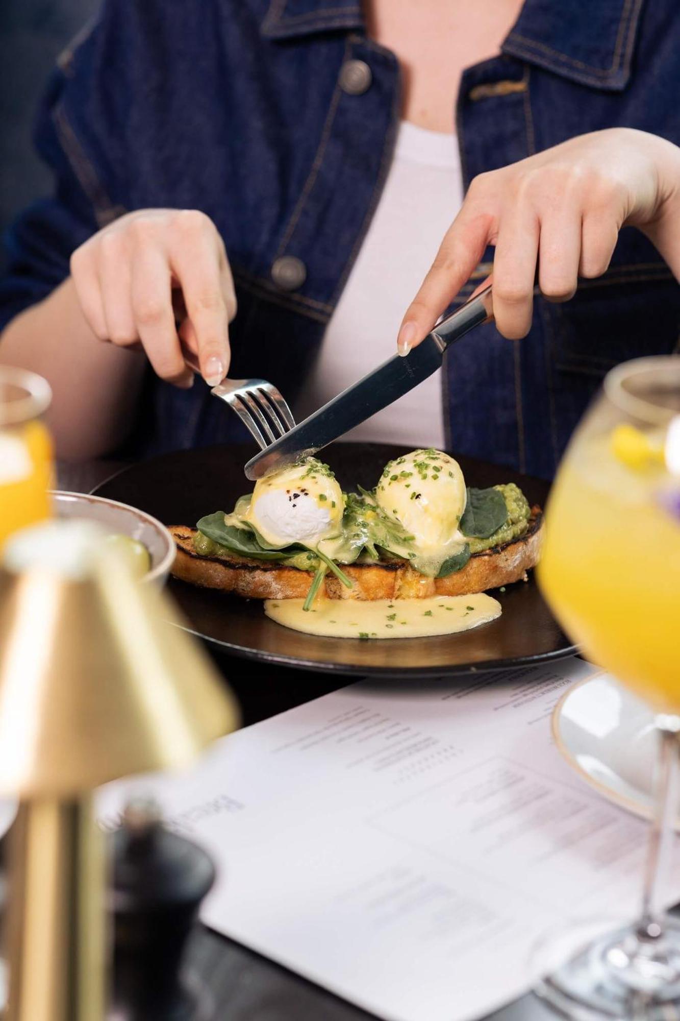Citysuites 2 Aparthotel Manchester Exteriér fotografie The photo shows a person preparing to eat a dish of eggs Benedict. They are holding a knife and fork, ready to cut into the meal, which features poached eggs on top of a slice of toast, accompanied by spinach and a creamy hollandaise sauce drizzled o