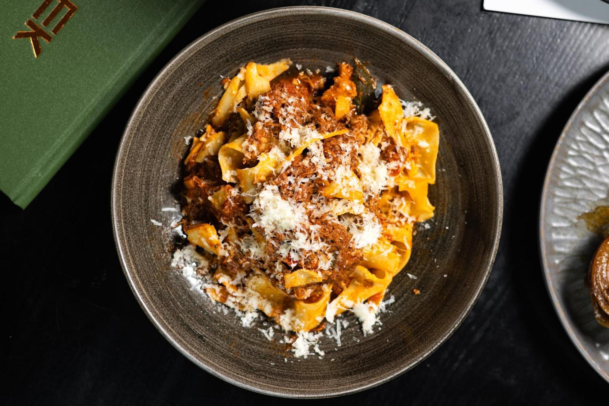 Citysuites 2 Aparthotel Manchester Exteriér fotografie The photo shows a bowl of pasta featuring wide, ribbon-like noodles, likely pappardelle, fully coated in a rich meat sauce. The dish is garnished with a generous sprinkling of grated cheese, adding a creamy texture to the presentation. The dark bowl 