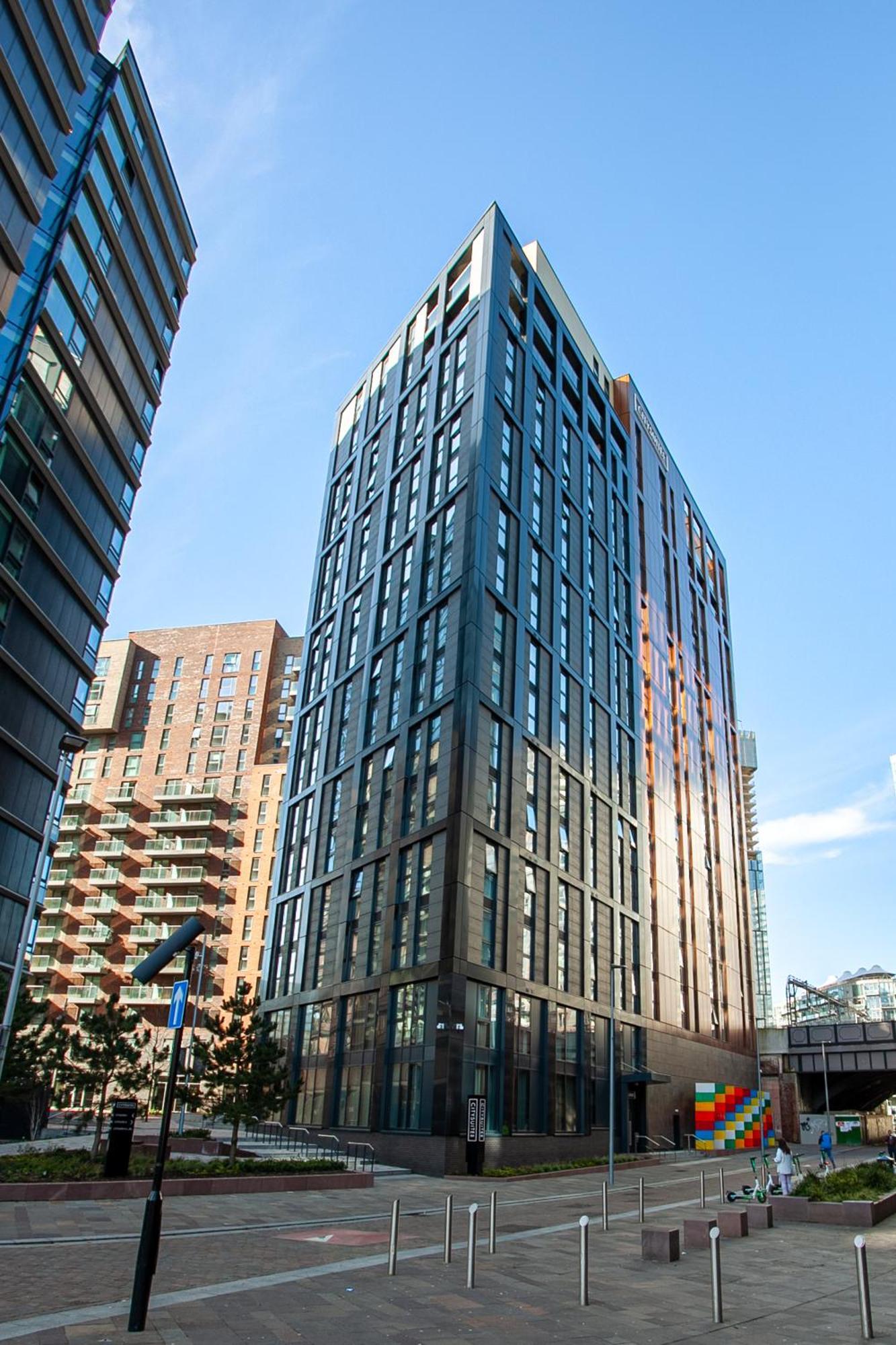 Citysuites 2 Aparthotel Manchester Exteriér fotografie The photo shows a modern high-rise building with a sleek, glass façade. It is set against a blue sky, indicating a clear day. The building appears to have multiple floors with large windows that reflect light. Nearby, there are additional buildings, 